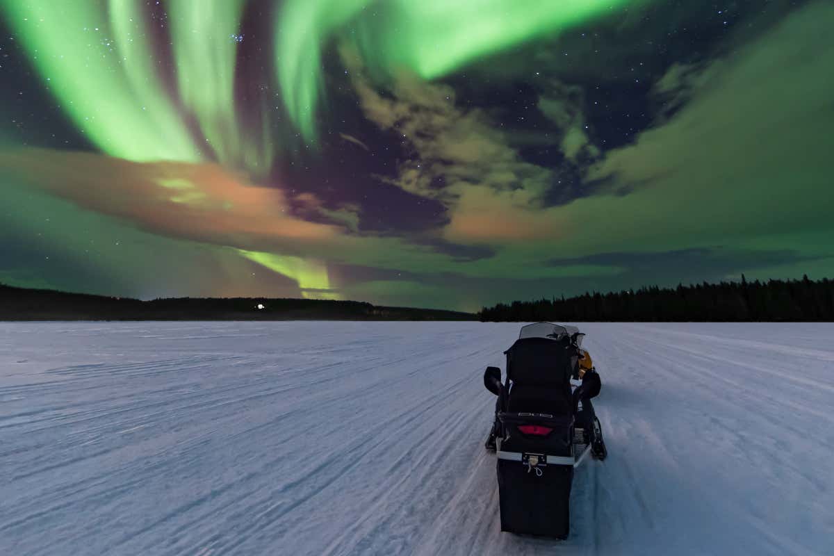 Una moto de nieve recorriendo una llanura por la noche bajo las luces verdosas de la aurora boreal