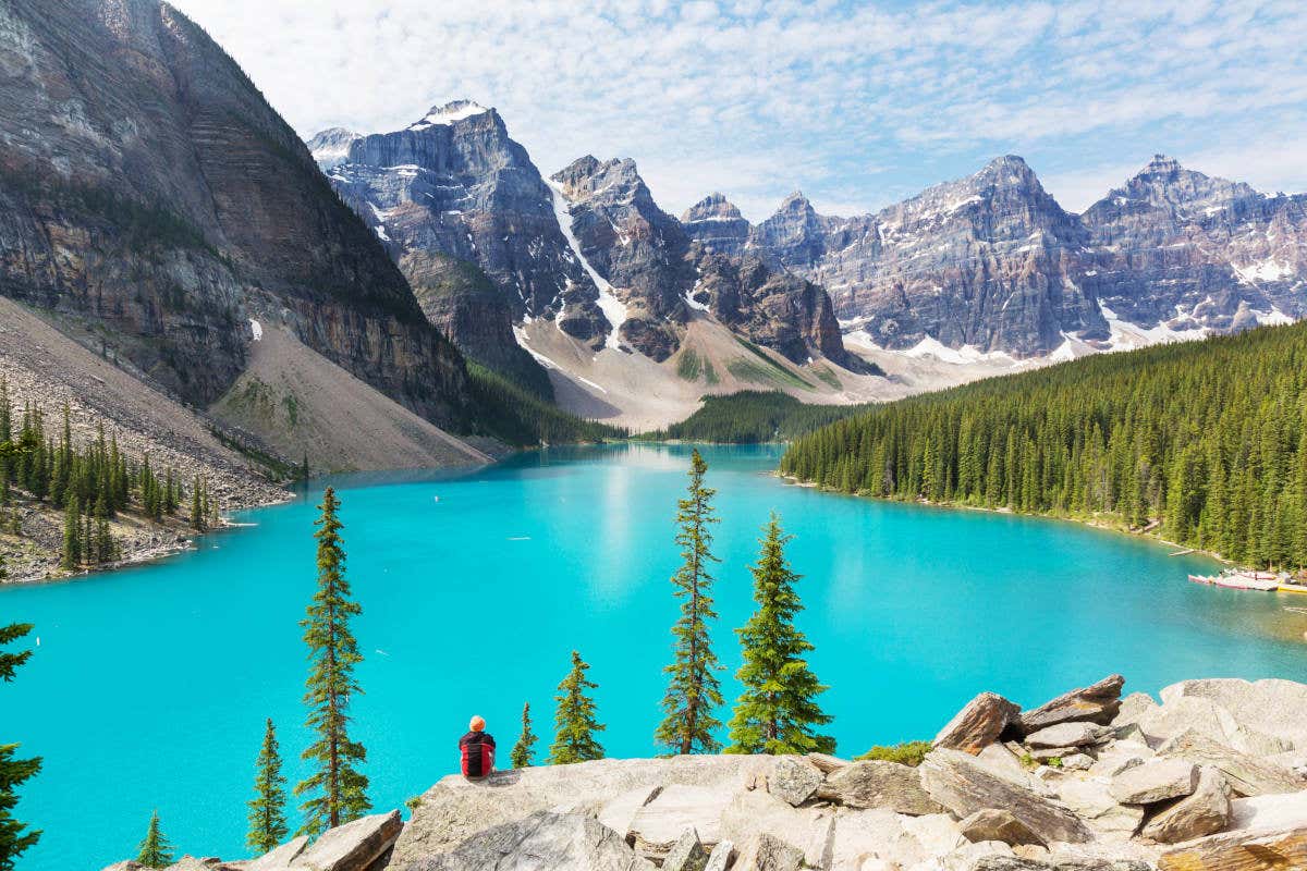 Escursionista di spalle seduto su una roccia davanti a un lago azzurro canadese circondato da alberi e montagne