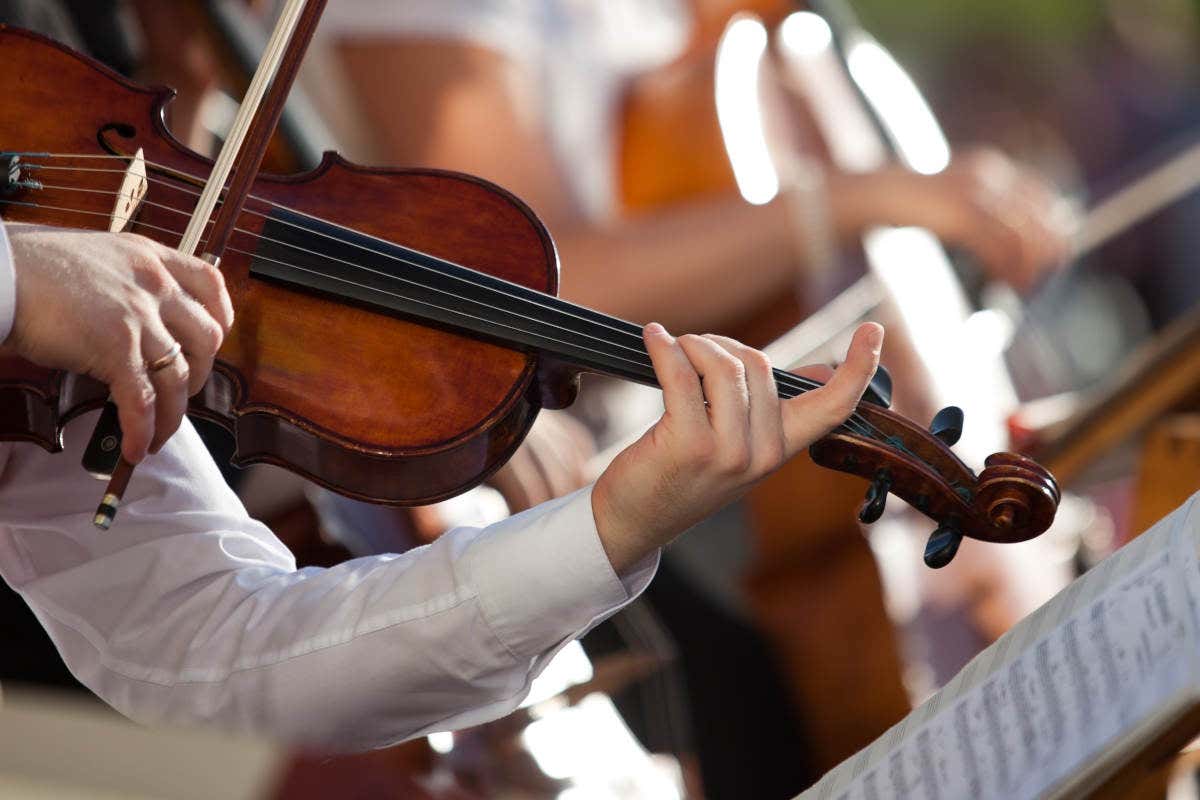 Un musicista di cui non è ripreso il volto, suonano un violino. Le braccia e le mani sembrano di un uomo che indossa una camicia bianca, sullo sfondo, fuori fuoco, si intravedono altri musicisti e un leggio con uno spartito