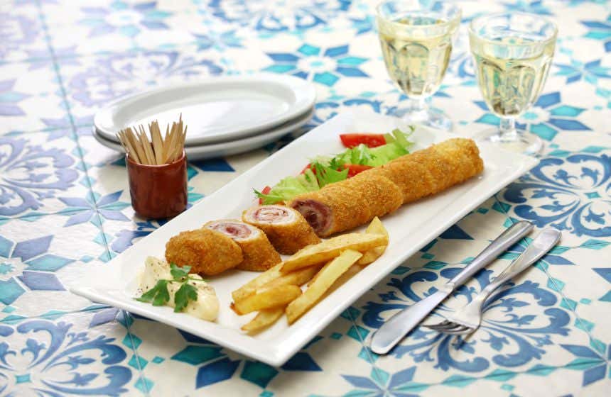 Um prato com flamenquín e batatas fritas, talheres ao lado, palitos e pratos de outro lado e duas taças ao fundo