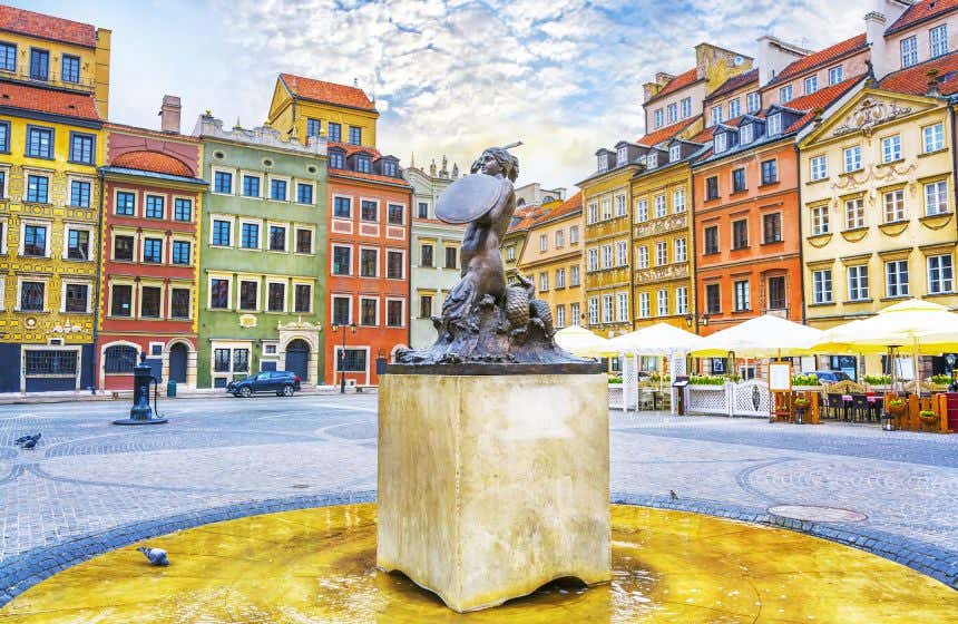 Monumento no centro de uma praça com diversos edifícios coloridos ao fundo e mesas de restaurantes ao lado