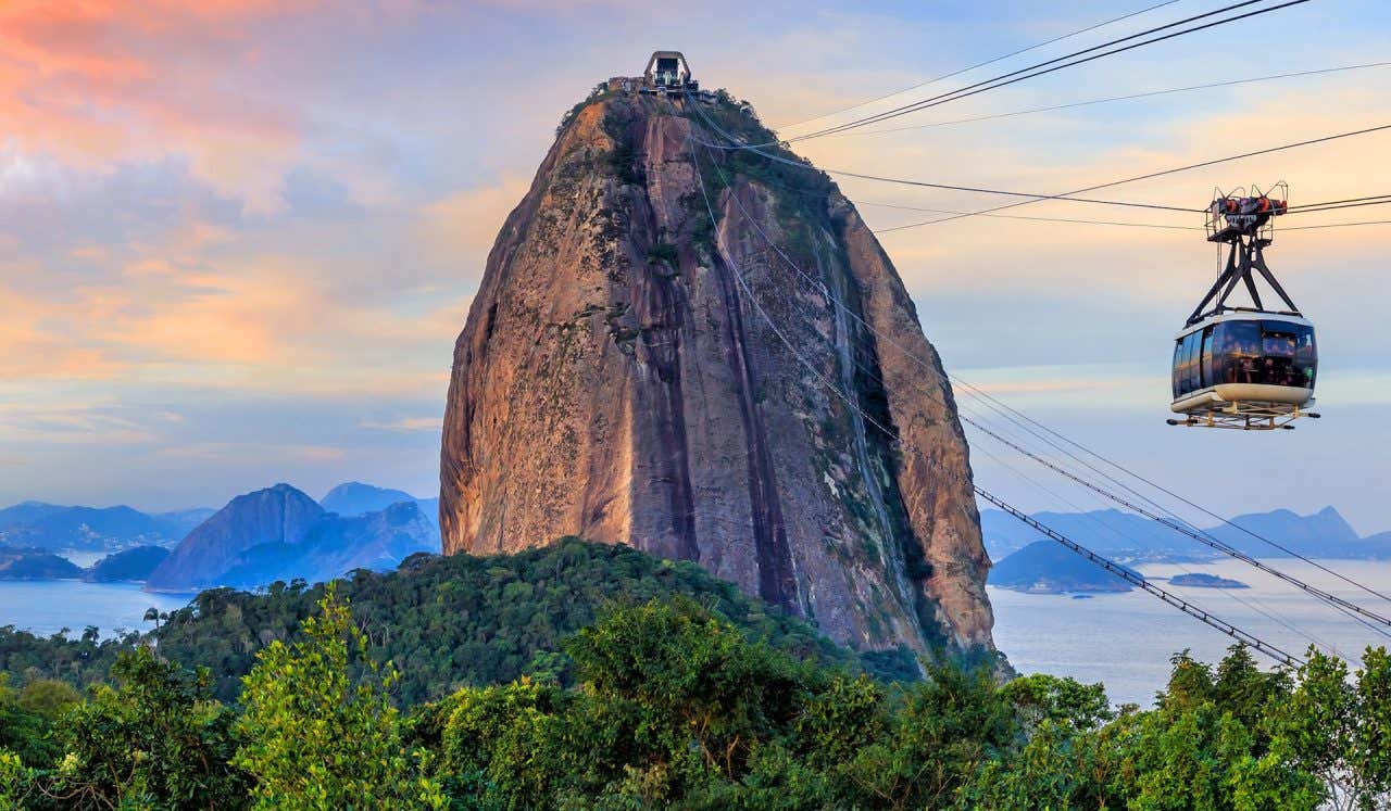 Il Pan di Zucchero a Rio de Janeiro, un enorme picco di granito che si alza sopra la vegetazione tropicale, illuminato dalla luce calda del tramonto. Sulla destra una cabina della funivia viaggia sospesa sui cavi, mentre sullo sfondo si vedono il mare e le montagne della baia di Guanabara sotto un cielo rosa e azzurro