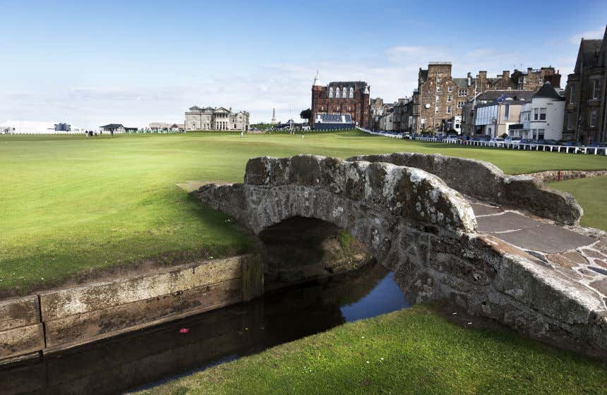 Gramado e pontes no campo de golf de Old Course de Saint Andrews com edifícios ao fundo.