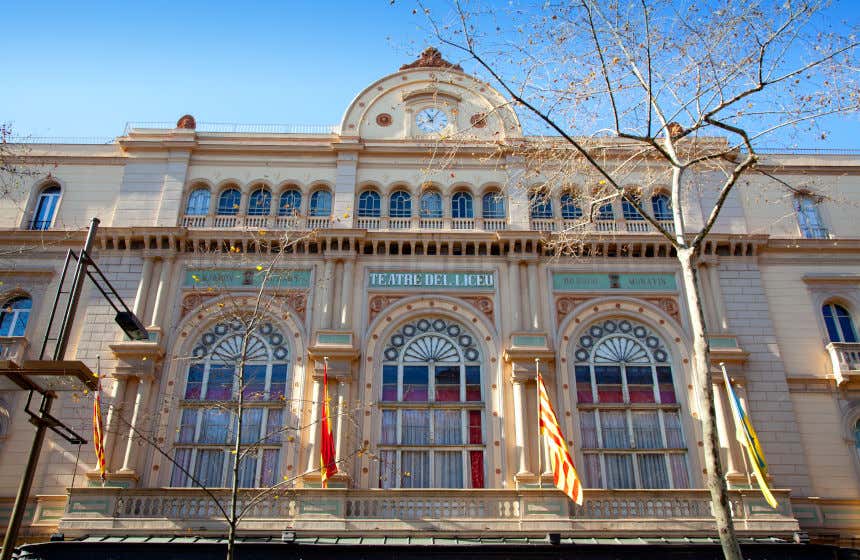 Vista frontal del Teatre del Liceu de Barcelona con las banderas de España y Cataluña colgando de su balconada y un árbol sin hojas en primer plano.