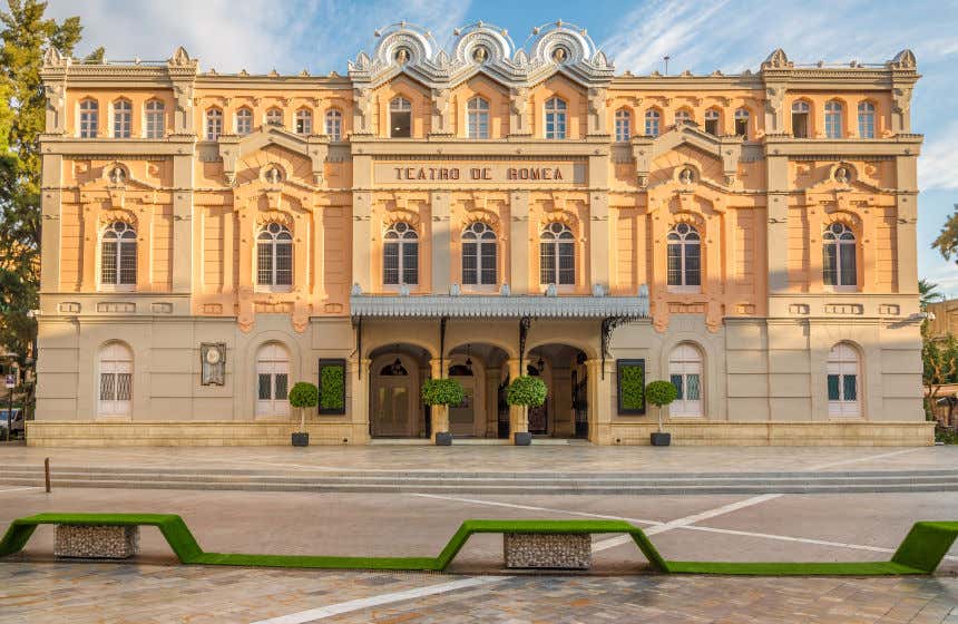 Vista frontal de la fachada gris y naranja pastel del teatro de Romea en Murcia con unos bancos en primer plano.