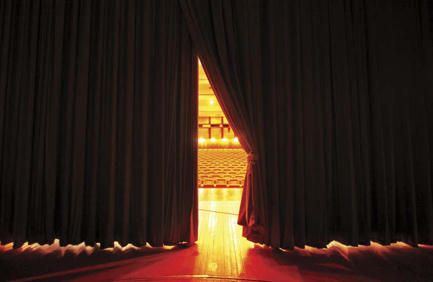 Vista del patio de butacas desde el escenario de un teatro con un gran telón rojo medio corrido.