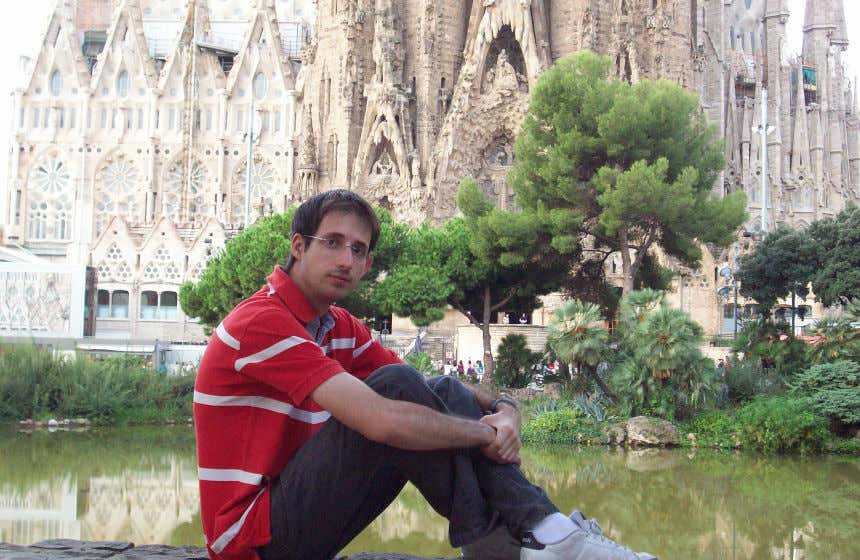 Javier Alameda sentado frente a un pequeño lago artificial rodeado de vegetación con el pórtico de la Sagrada Familia de Gaudí al fondo.