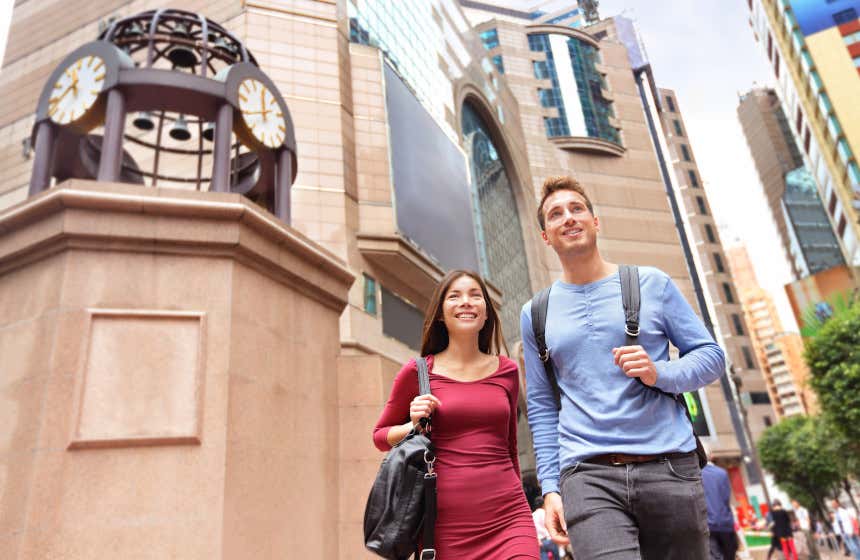 Una pareja de jóvenes paseando por Causeway Bay, el distrito comercial más popular de Hong Kong. Se puede divisar al fondo varios edificios y un reloj con cuatro esferas y campanas.