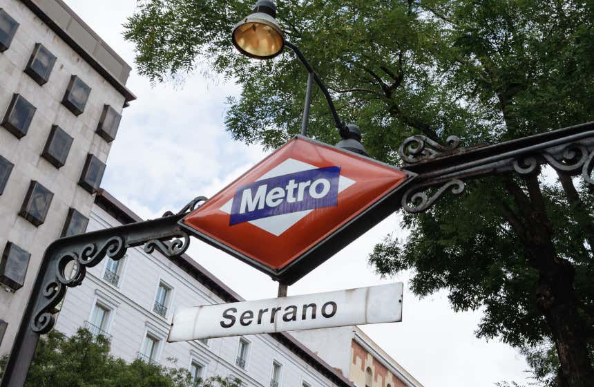 Cartel identificativo del acceso a una de las bocas de metro de la estación de Serrano, en el centro de Madrid. Tras la señal, se pueden ver algunos edificios y un gran árbol.