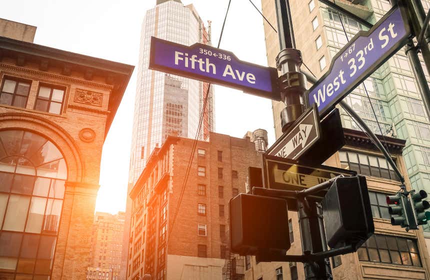 Carteles azules en un semáforo del centro de Manhattan indicando cómo llegar a la Quinta Avenida de Nueva York. Al fondo se pueden ver altos rascacielos bajo la luz del atardecer.