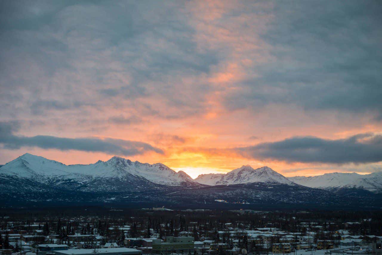 A wide angle panoramic view of a quiet snow covered town, snow capped moutains in the distance, with a brillant sunset setting over the horizon