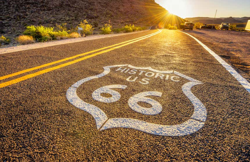 Vista del asfalto de la Ruta 66 con un cartel identificativo de esta histórica carretera. Al fondo se pueden ver varios coches y una puesta de sol.