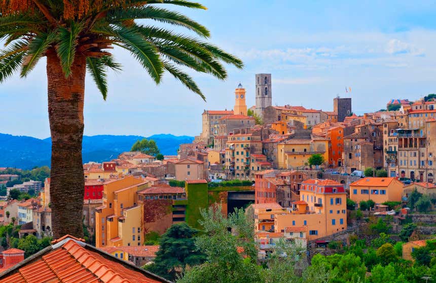 Vista panorámica del pueblo francés de Grasse con una palmera a la izquierda y numerosas casas coloridas y algunas torres a la derecha.