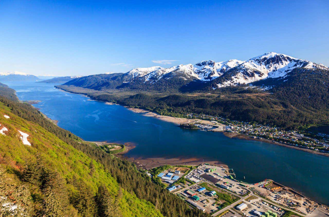 Aerial panoramic view of a waterfront valley, snowy mountains on the right side of the river, a lush green mountain on the left side