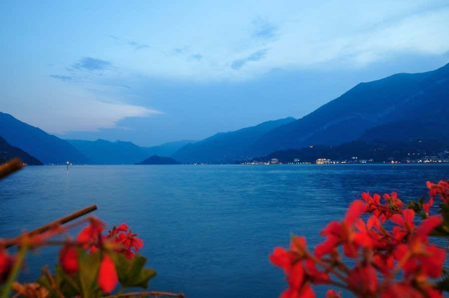 Lago di Como al calar della sera, con fiori rossi a destra e a sinistra e le montagne a fare da sfondo.