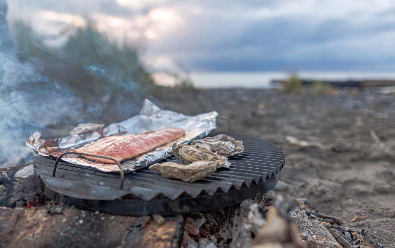 A cut of salmon sits on a piece of foil, placed on a grill plate over a fire in the middle of the woods, smoke rising above
