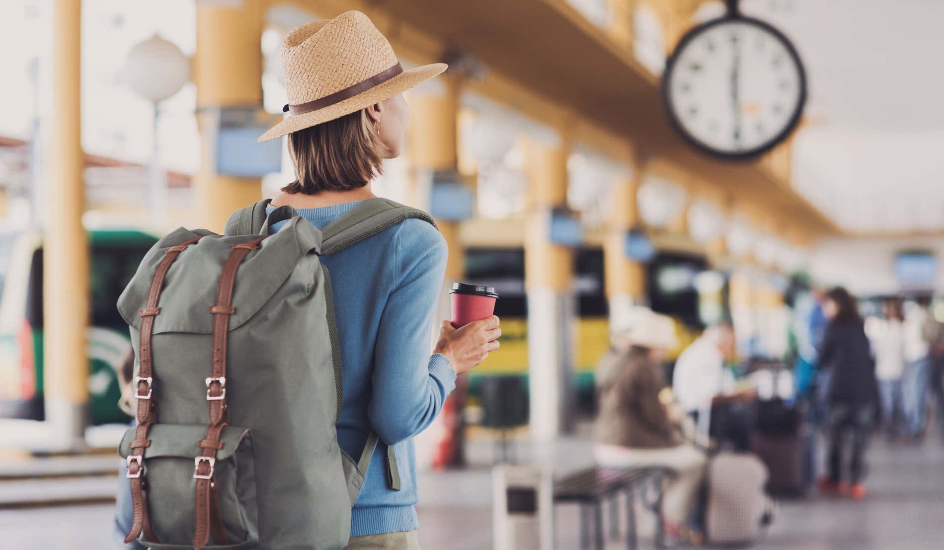 Uma mulher com uma mochila e café na mão olhando para um relógio em uma estação de ônibus internacional