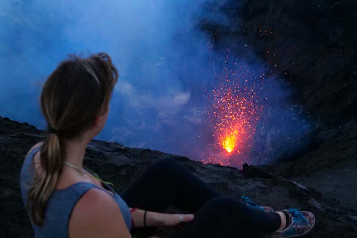 Uma mulher com roupa esportiva sentada de costas olhando a lava brotando da cratera do Monte Yasur, em Vanuatu