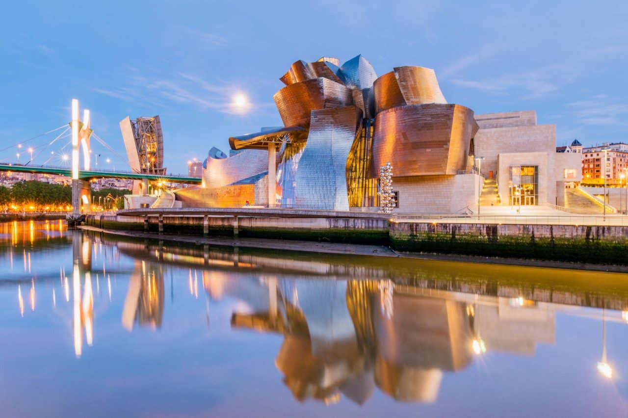 A wide shot of the exterior of the Guggenheim Museum Bilbao in the evening, with its reflection visible on the water below