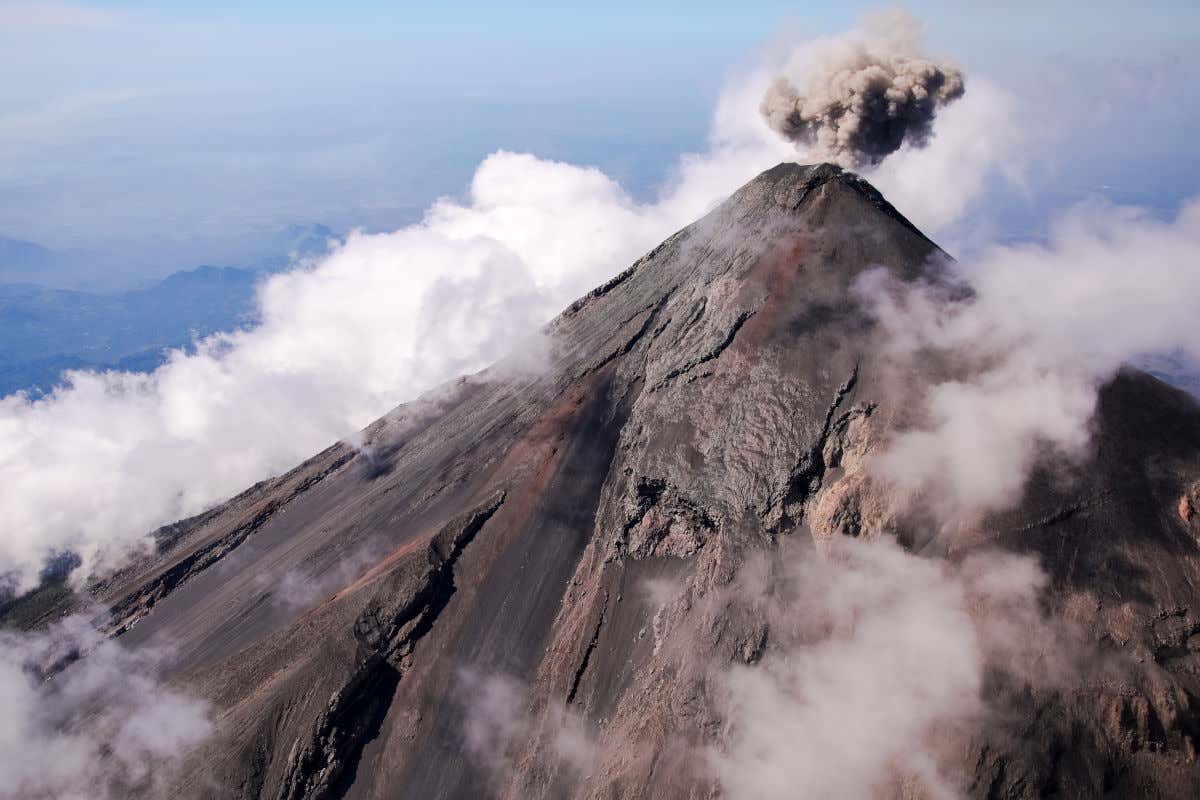 A cone shaped volcano emitting grey smoke from its peak and surrounded by white clouds