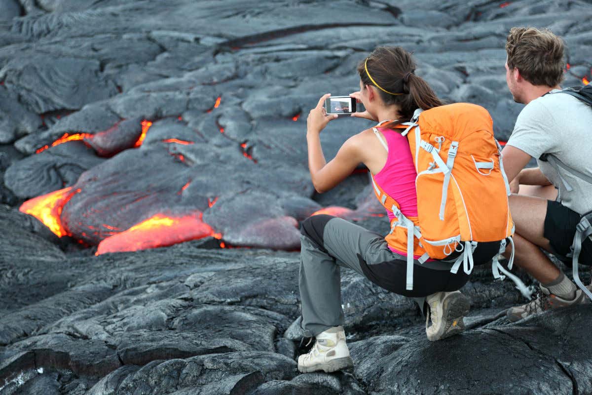 Dois turistas de costas com roupa esportiva tirando fotos dos fluxos de lava do Kīlauea, no Havaí