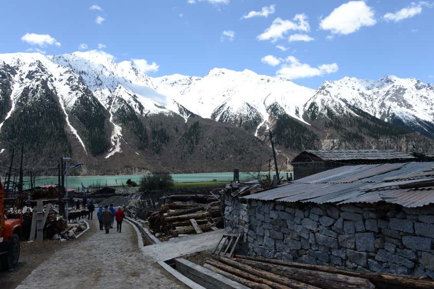 Motu, Tibet, with people walking along a path leading to a lake and a snow-covered mountain range under a blue sky