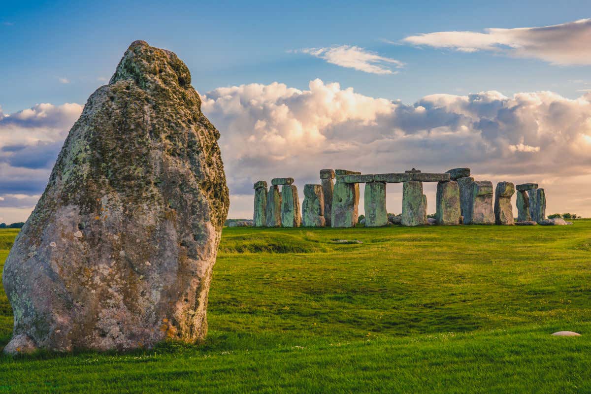 Una piedra frente al conjunto rupestre de Stonehenge bajo una capa de nubes y los colores del atardecer.