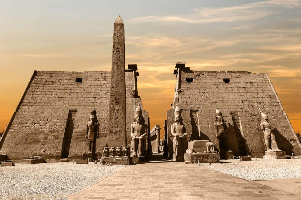 Vista frontal de la entrada del templo de Luxor con un gran obelisco y cinco grandes esculturas de piedra. Al fondo se pueden ver las altas columnas aun en pie y los colores del atardecer.