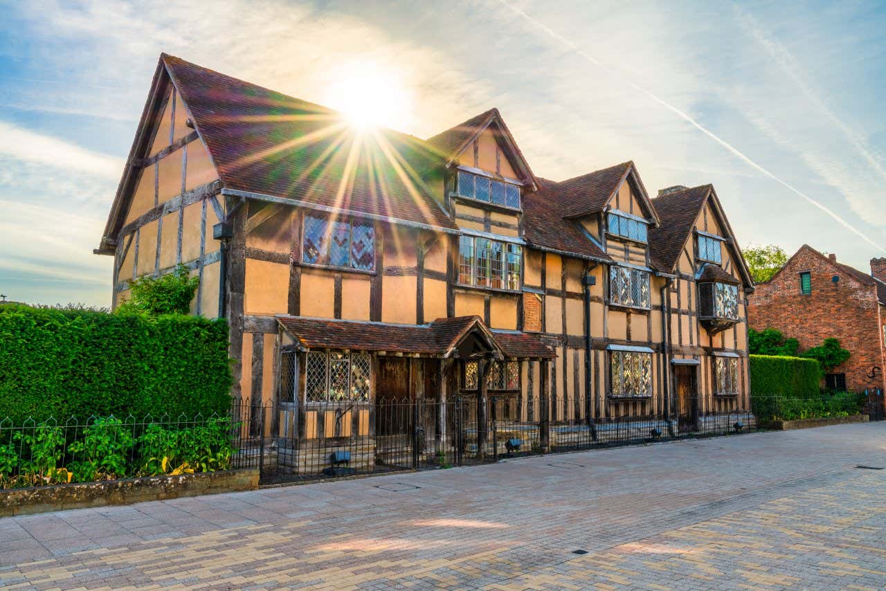A picture of Stratford-upon-Avon, Shakespeare's birthplace, with the sun shining above the house and beautiful green bushes either side.