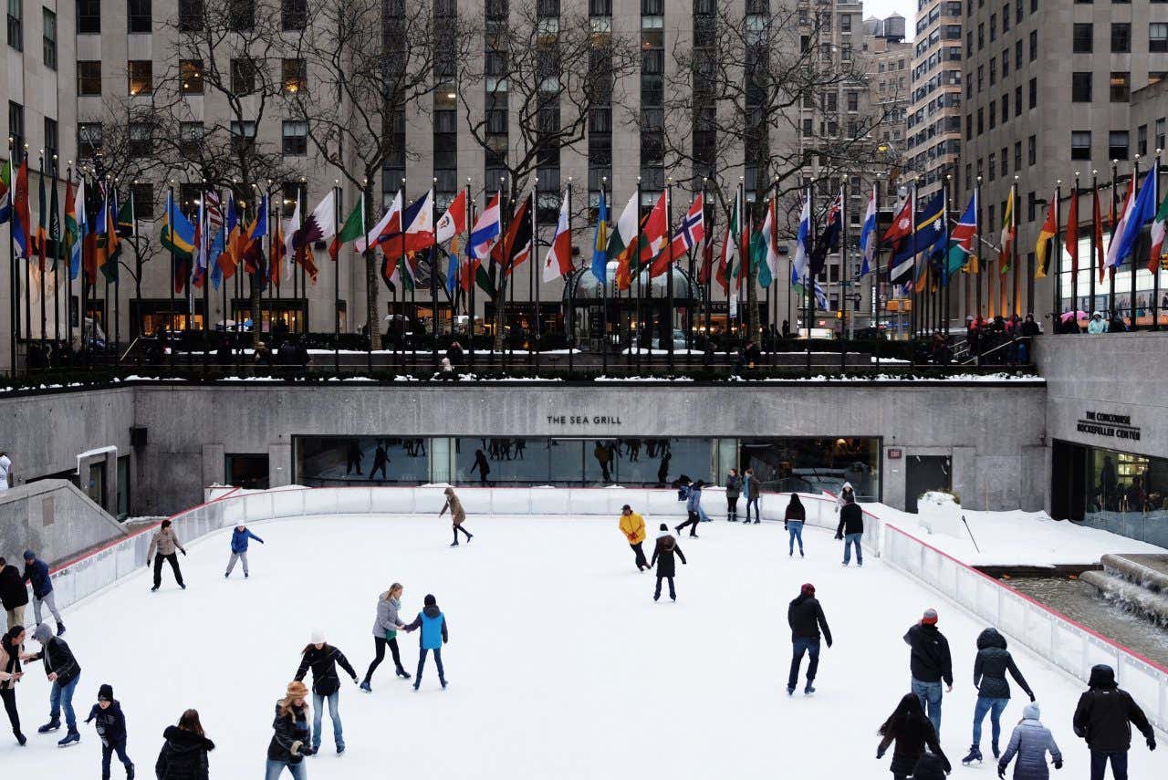 The Rink at Rockefeller Center in New York City, full of people ice skating, with all of the flags flying next to the rink.