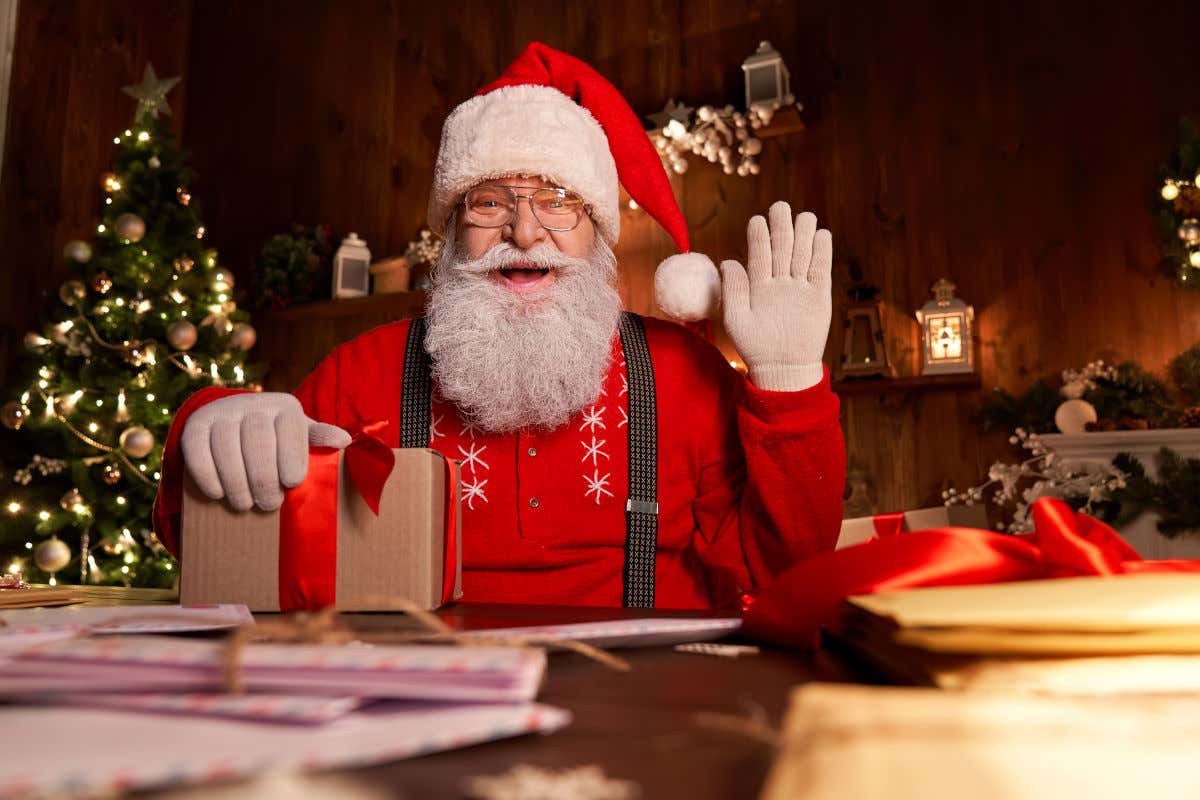 Papá Noel sentado y ataviado con un traje y gorro rojo mientras sujeta un regalo sobre una mesa y saludo al espectador. Al fondo se puede divisar un árbol de Navidad y varios farolillos.