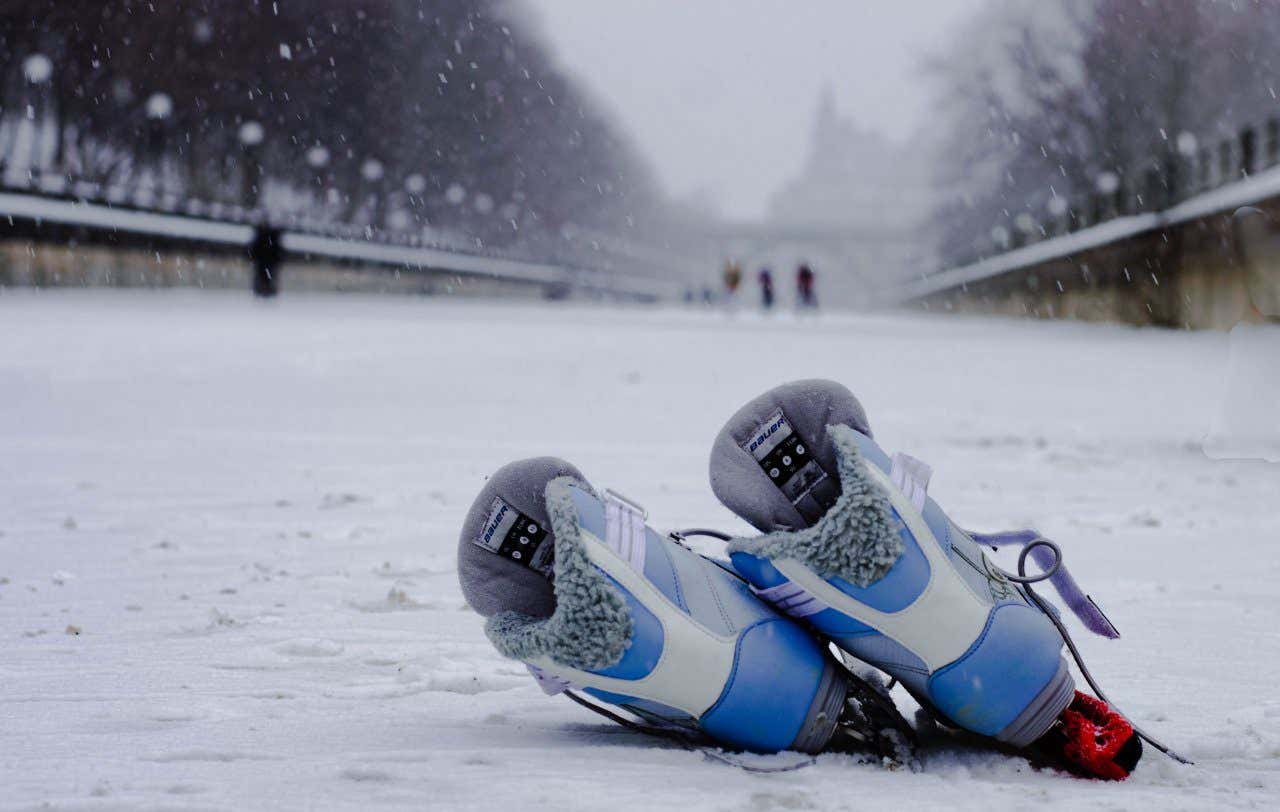 A pair of blue ice skates leaning over on Rideau Canal Skateway, with snow falling all around them and people ice skating in the distance out of focus.
