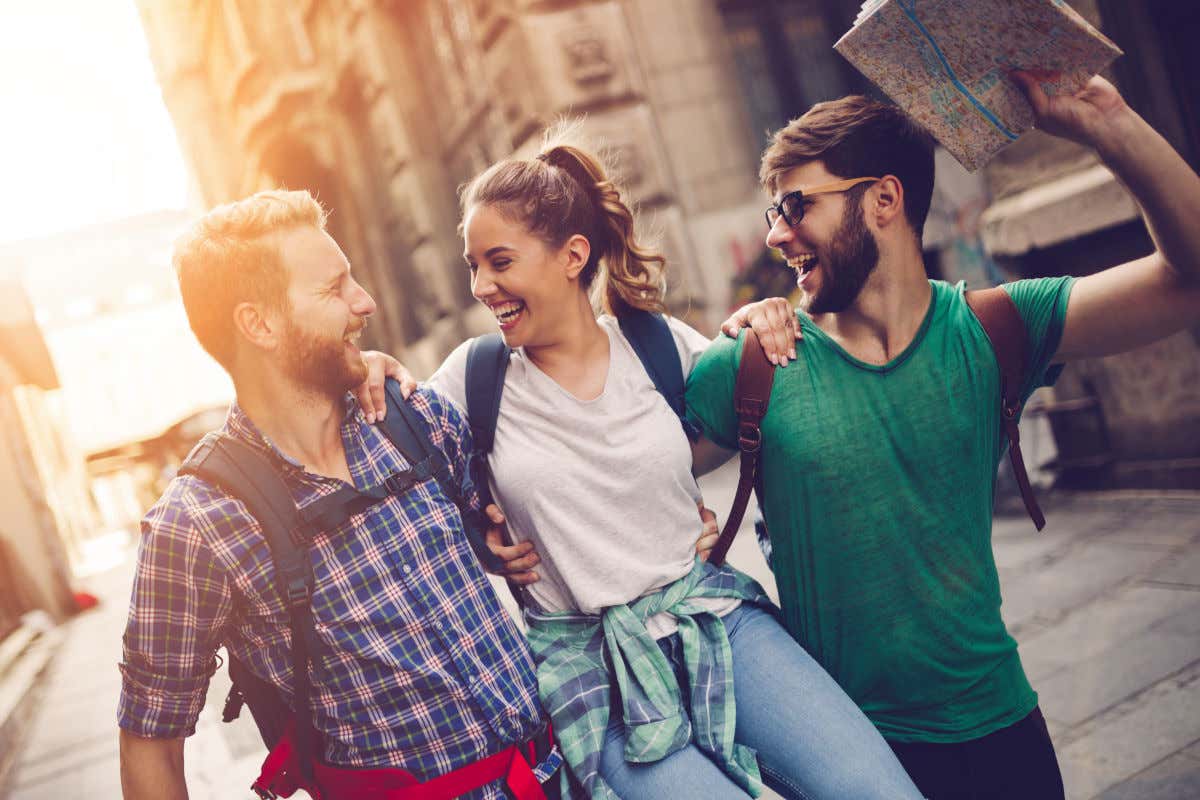 Un grupo de tres amigos, dos hombres y una mujer, con un mapa en mano disfrutando mientras hacen turismo por una calle soleada.