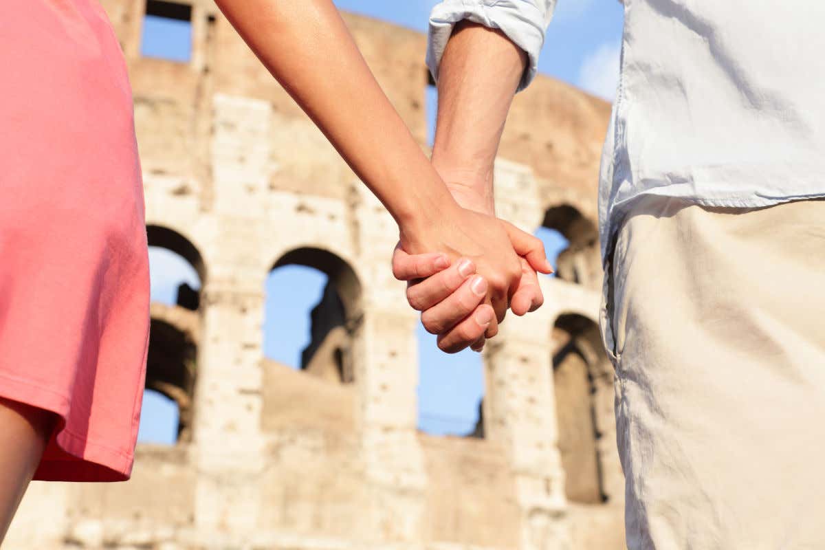 Dos manos entrelazadas de un hombre y una mujer frente al Coliseo de Roma.