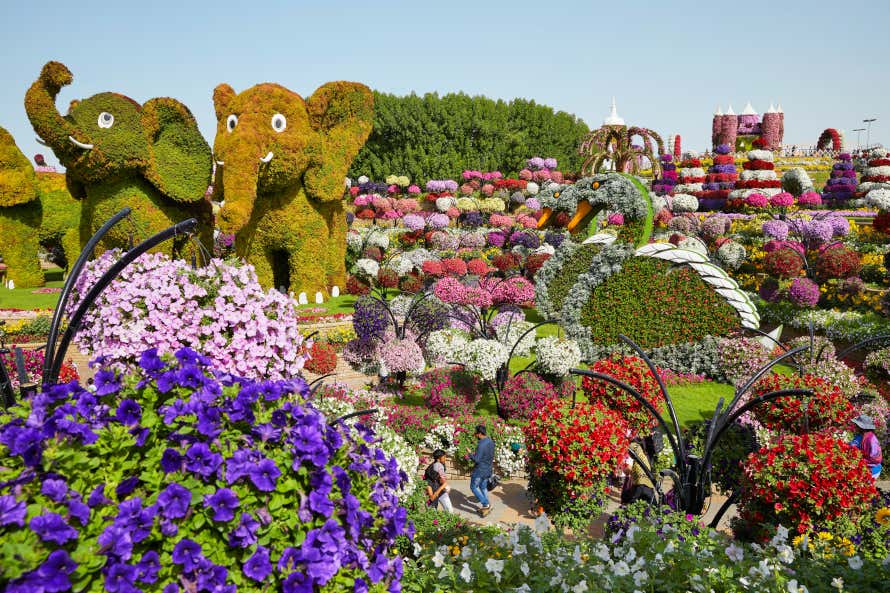 Pessoas caminhando em um jardim coberto de flores, árvores e até mesmo elefantes feitos de plantas