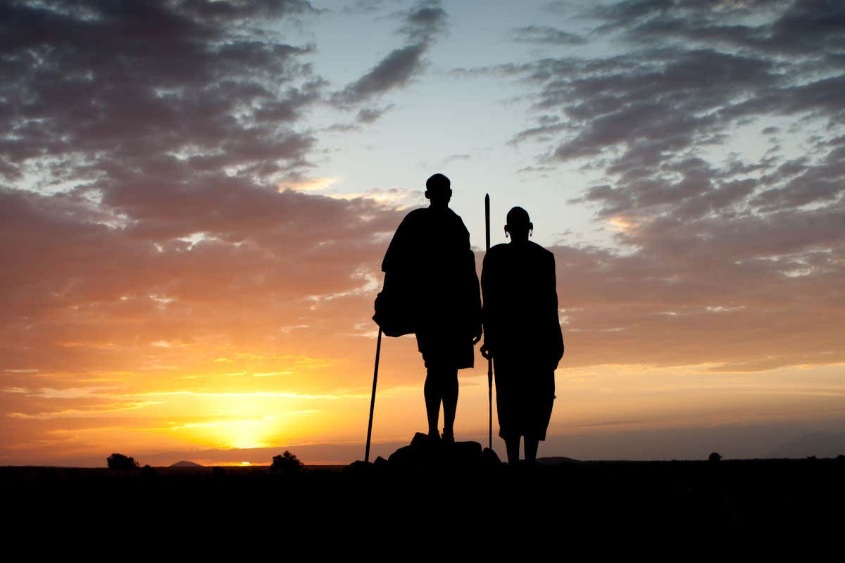 Dois membros de uma tribo de costas para a câmera contemplam o nascer do sol na zona de Masai Mara, no Quênia