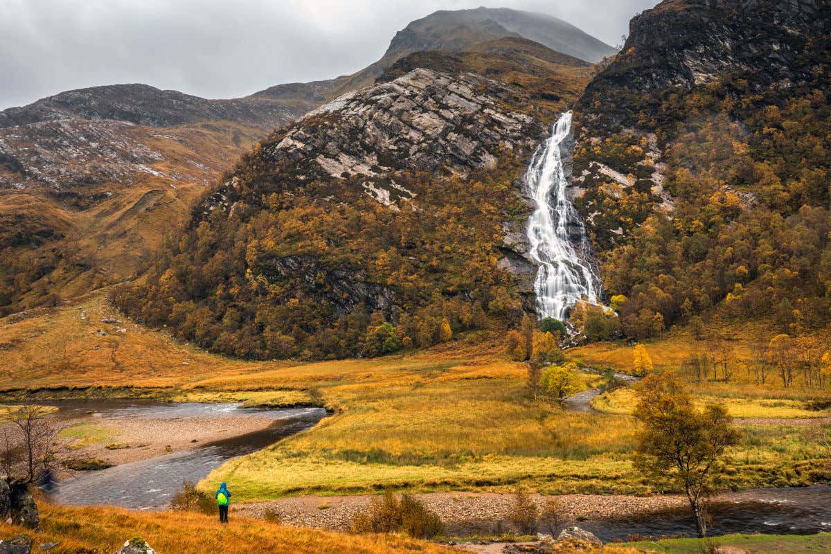 As cataratas de Steall fluindo pelas montanhas em um dia nublado e possivelmente de outono devido aos tons ocres e amarelos da vegetação