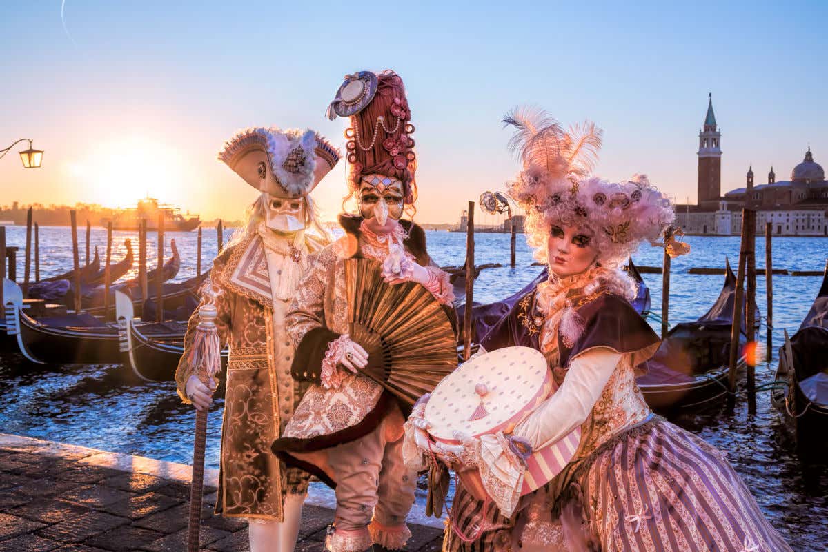 Disfraces y máscaras típicos del carnaval de Venecia frente a un embarcadero de góndolas con la plaza de San Marcos al fondo.