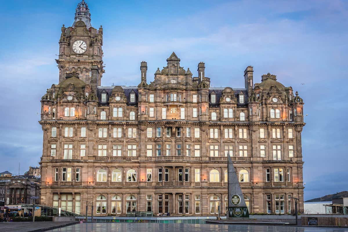 Fachada do hotel Balmoral de Edimburgo ao entardecer com as luzes exteriores acesas e sua típica torre de relógio marcando as quatro da tarde