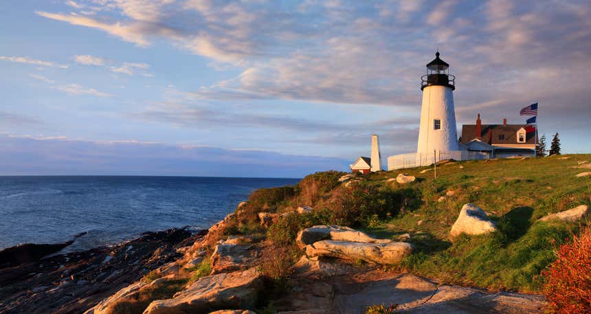 Pemaquid Point Lighthouse ao entardecer com uma casa ao fundo com a bandeira dos Estados Unidos