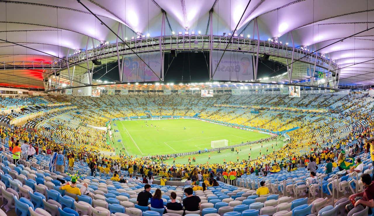 L’interno dello stadio Maracanã di Rio de Janeiro, illuminato di sera, con il campo verde al centro e la copertura bianca sopra gli spalti. Sulle tribune, tra seggiolini bianchi e blu, si vedono molti tifosi con le maglie gialle del Brasile e qualche bandiera