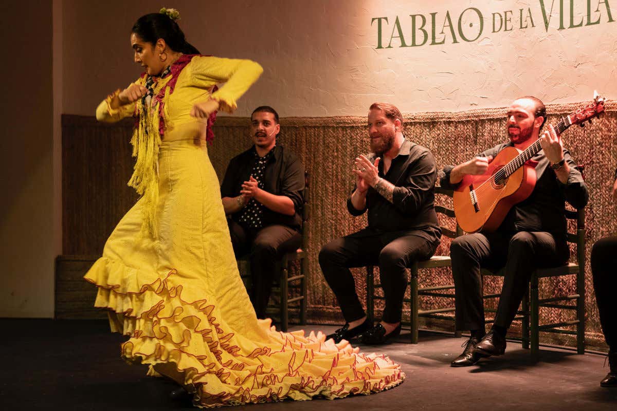 Une danseuse de flamenco avec une robe à queue jaune devant trois musiciens tapant des mains et jouant de la guitare