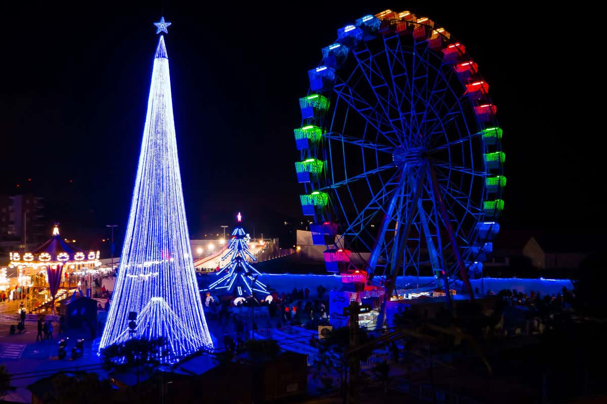 Vista nocturna de una gigantesca noria, junto a dos árboles de Navidad artificiales y un carrusel en la feria de Navidad de Torrejón de Ardoz