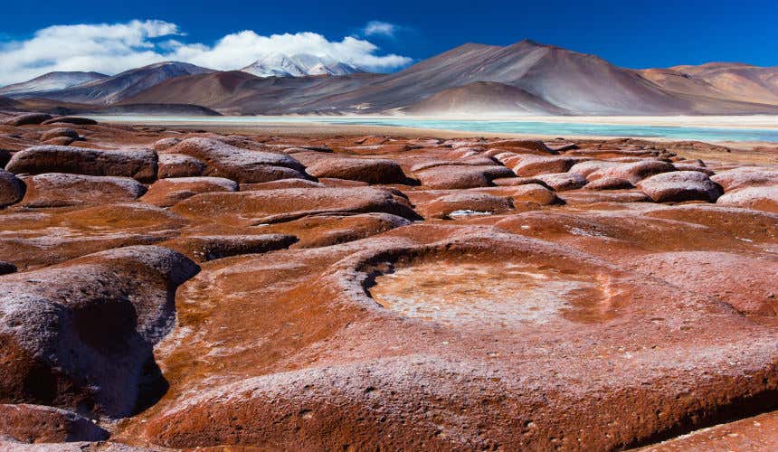 Brown smooth rocks by a light blue lake and smooth mountains in the background in the Atacama desert