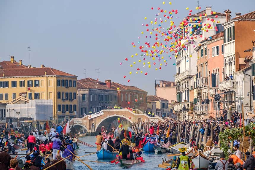 People in costume strolling along a Venetian canal in boats while the sky is filled with balloons