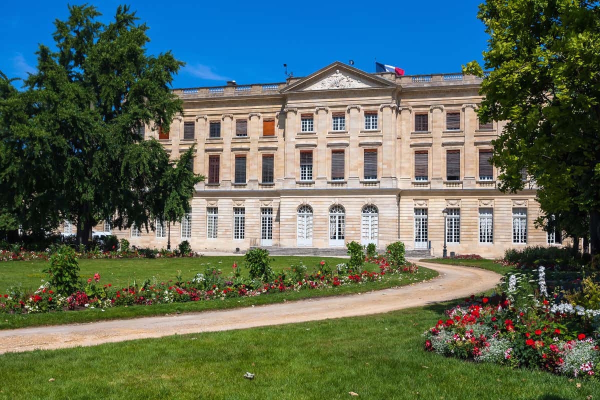 Fachada do Museu de Belas Artes de Bordeaux em frente a um jardim com a bandeira francesa em movimento.