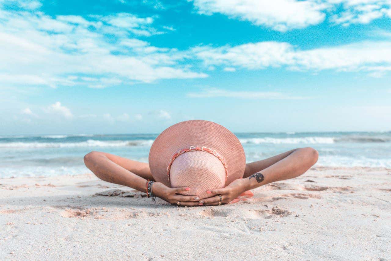 Woman's head with a sunhat on and her arms up above as she lays on the white sand beach of Playa del Carmen in front of the Caribbean Sea in Mexico