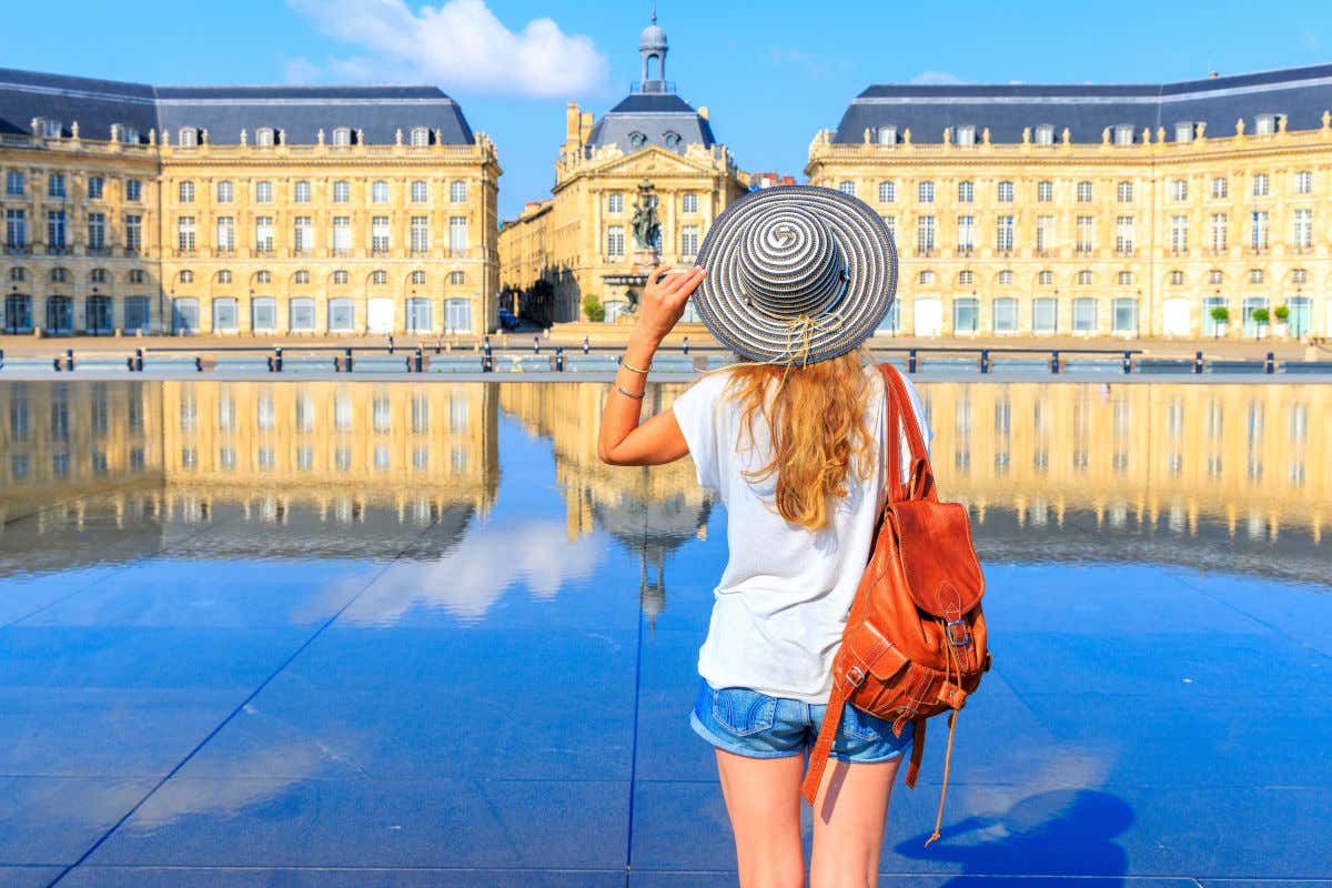 Uma jovem com roupas de verão olhando para um edifício de Bordeaux refletido no chão molhado.