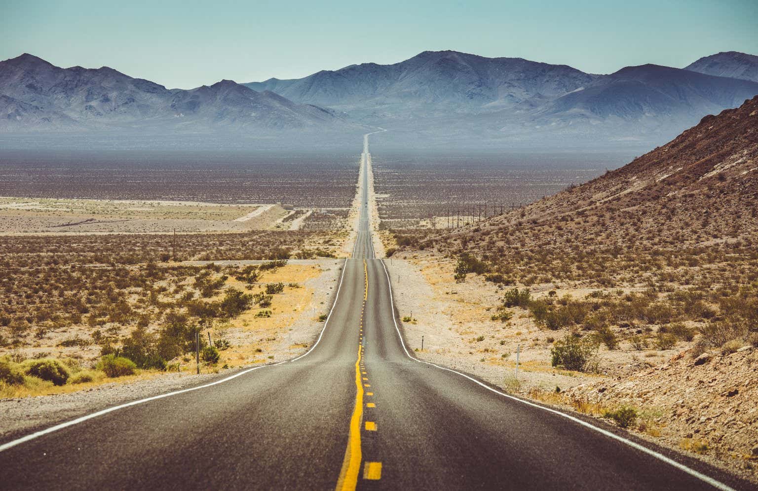 Uno scorcio di strada che attraversa una vasta pianura statunitense e si perde all'infinito, verso le montagne