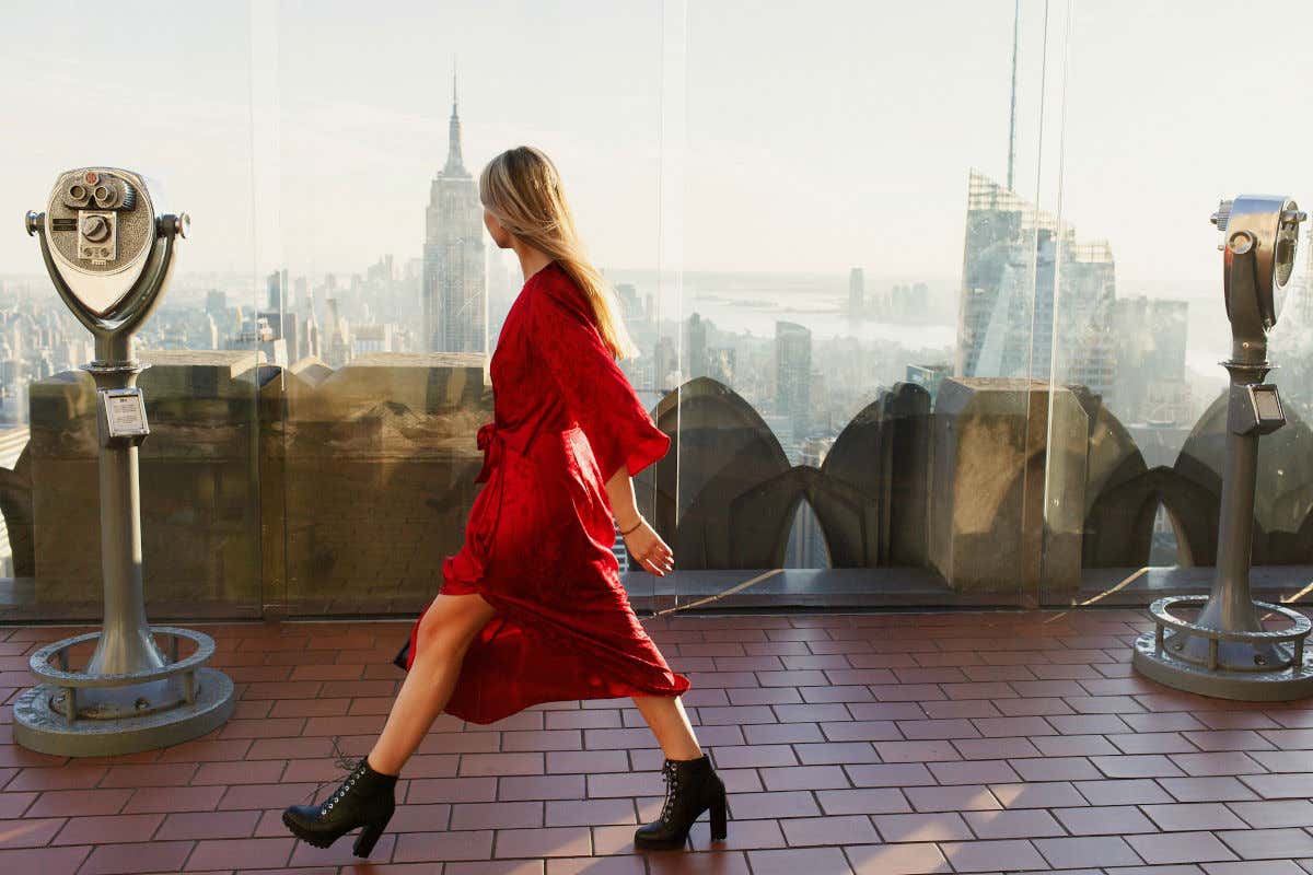 Una mujer con vestido rojo y botas negras caminando frente a las cristaleras del Top of the Rock, uno de los mejores miradores de Nueva York